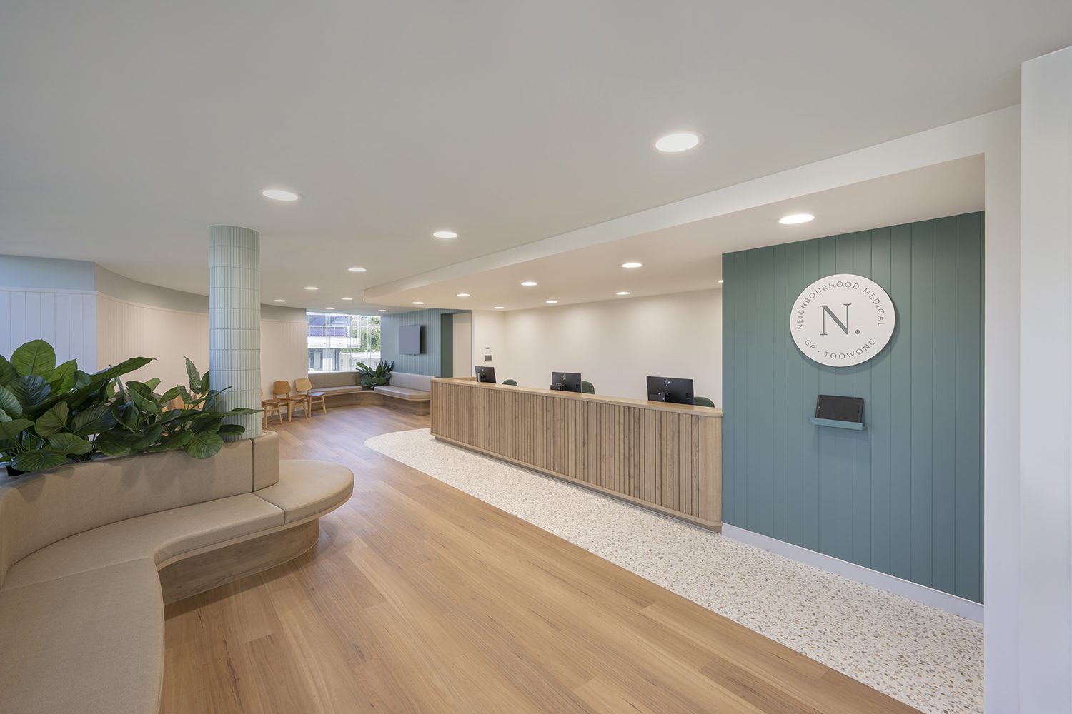 Reception with a curved timber counter, a sage green wall carrying a round clock, planting and a waiting bench on a timber floor